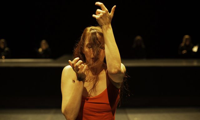 Woman in red dress with long hair covering her face stands with closed eyes and arms lifted in a gesture