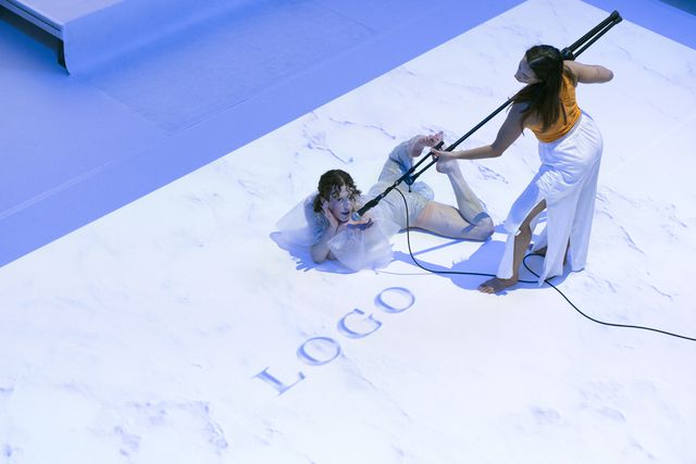Performer lying on the floor, smiling into the camera while another performer holds a microphone for them