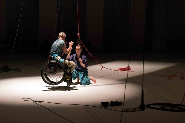 Performer with rope knotted into her hair pulls up another performer with wheelchair